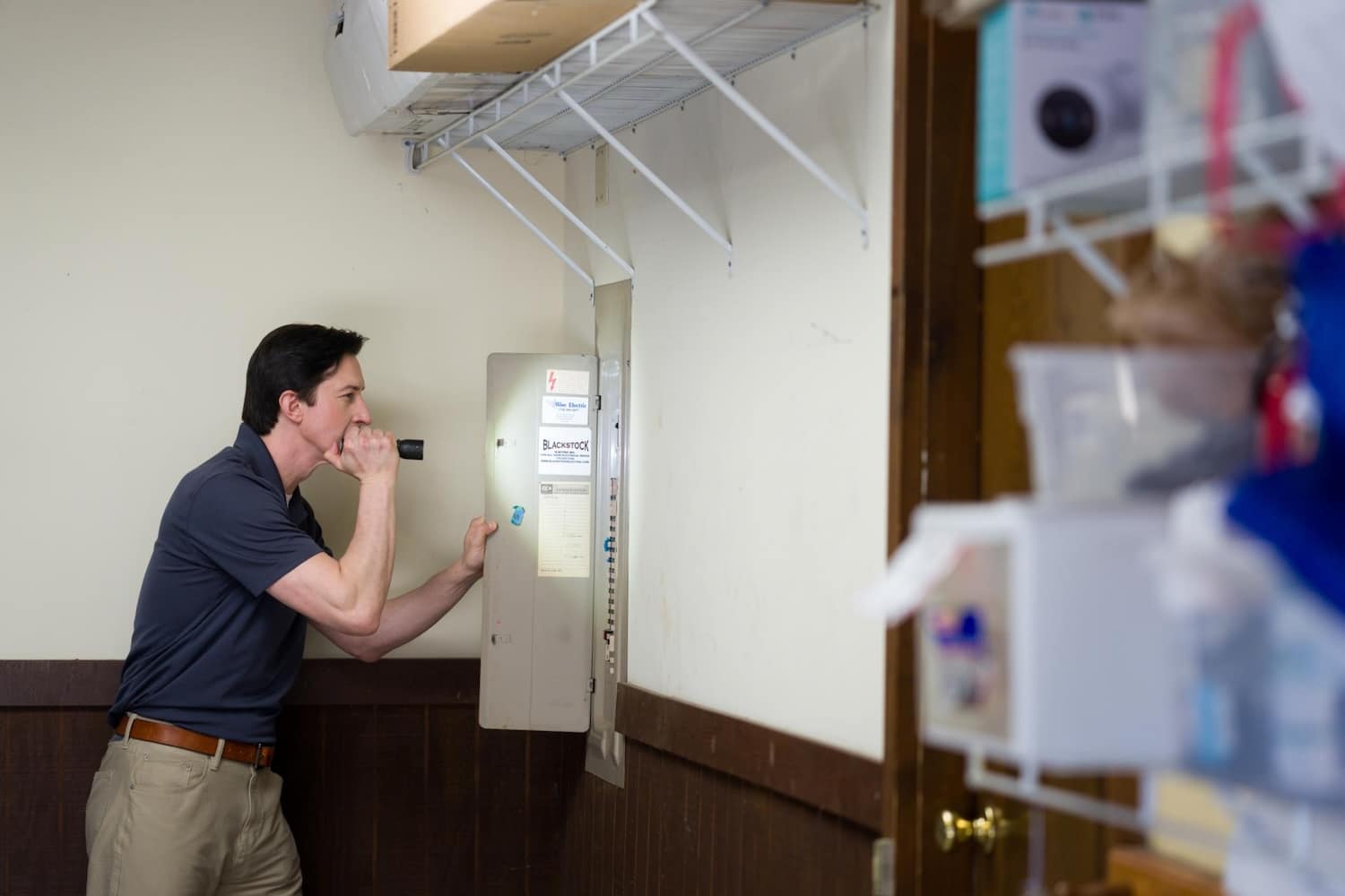 A man shines a flashlight into an open electrical breaker panel mounted on a wall.